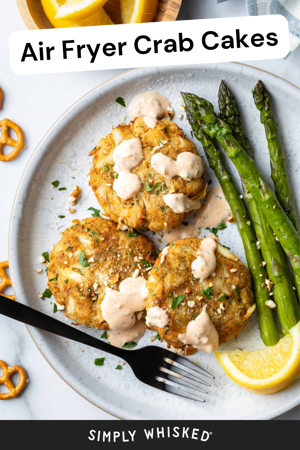 A plate with two air fryer crab cakes topped with sauce, grilled asparagus, a lemon wedge, and a black fork. Air Fryer Crab Cakes text is at the top, and Simply Whisked is at the bottom.