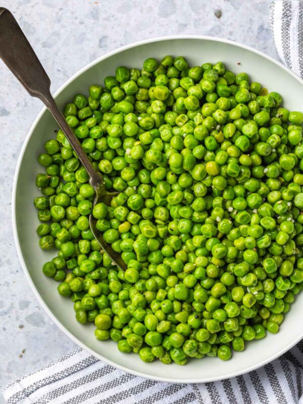 A bowl filled with bright green peas, cooked just right and ready to eat, with a spoon resting inside. The bowl sits on a light-colored surface next to a striped cloth—perfect for learning how to cook frozen peas deliciously.