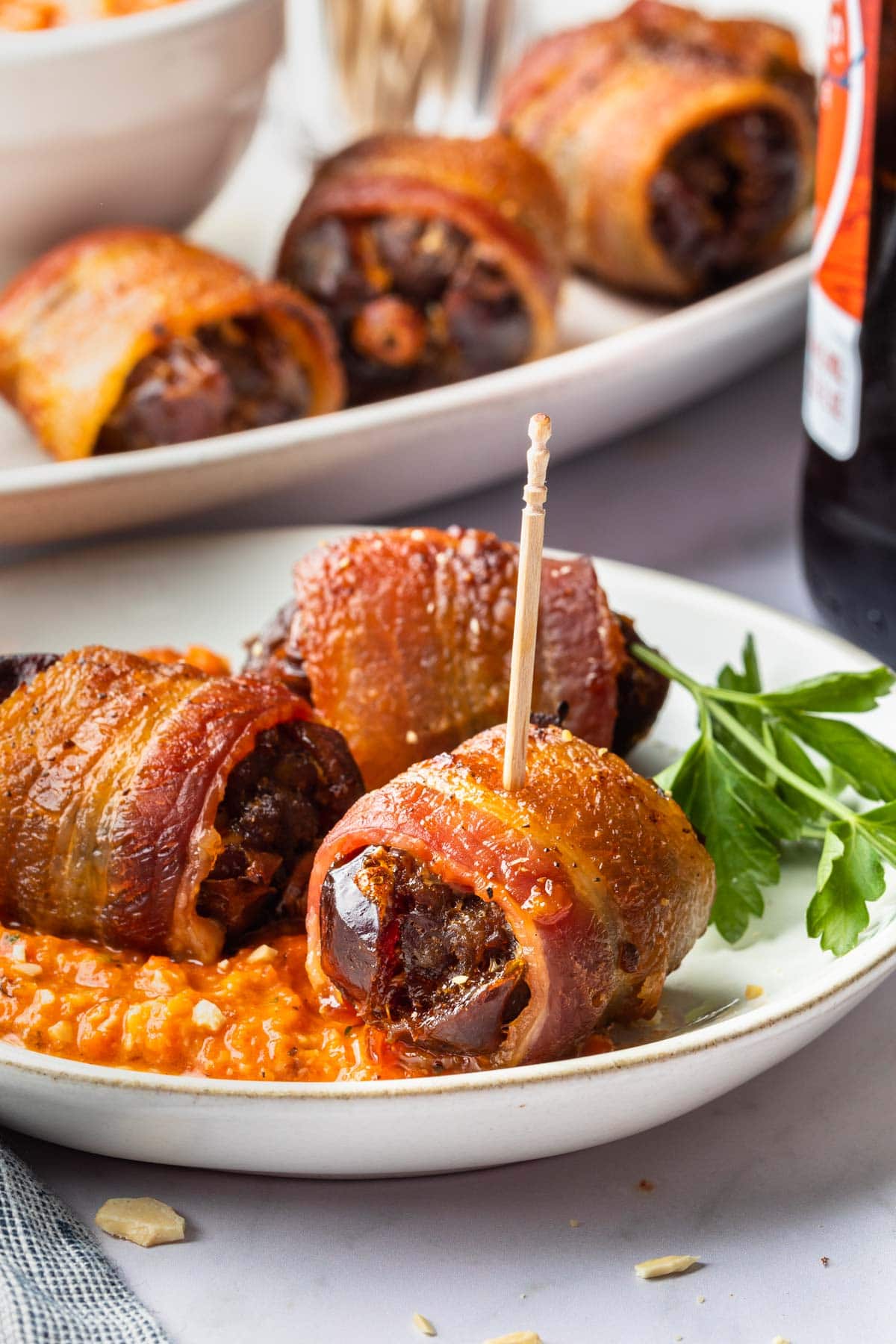 Chorizo stuffed dates on an appetizer plate with a serving tray of them in the background. 