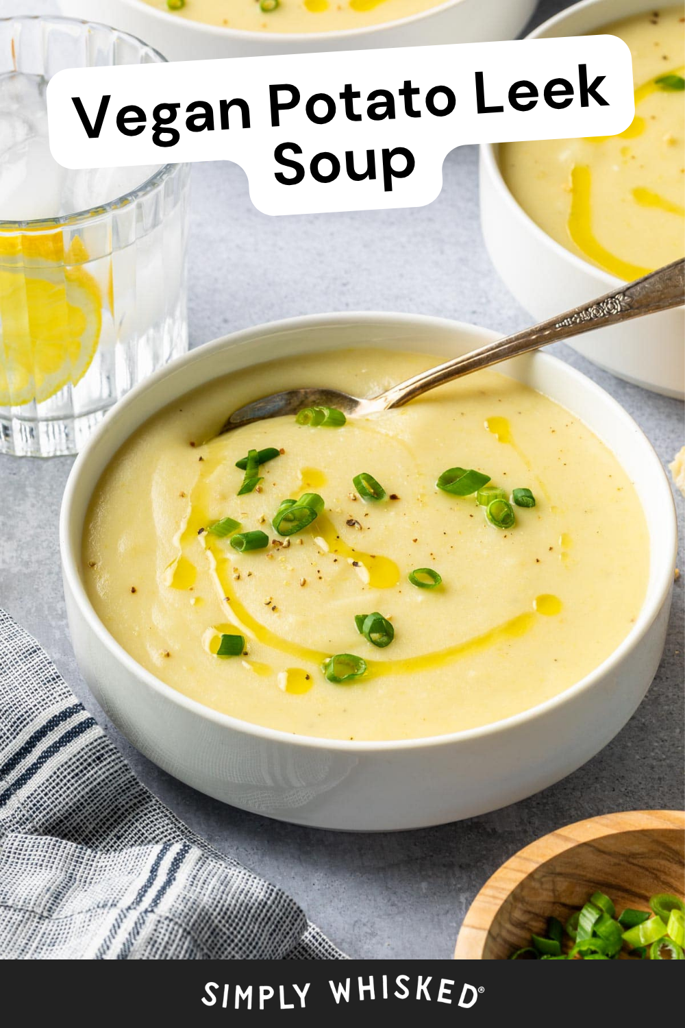 A bowl of creamy Vegan Potato Leek Soup topped with chopped green onions and a drizzle of oil, with a spoon nestled inside. A glass of water with lemon and a striped napkin sit beside the bowl, inviting you to enjoy this comforting meal.