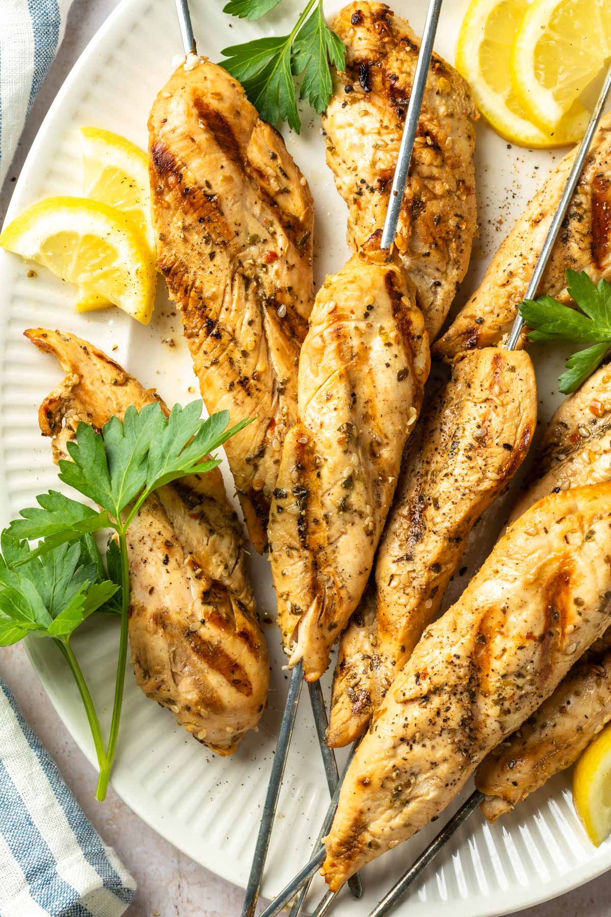 Grilled chicken tenders on metal skewers on a serving platter with lemon slices and sprigs of fresh parsley. 