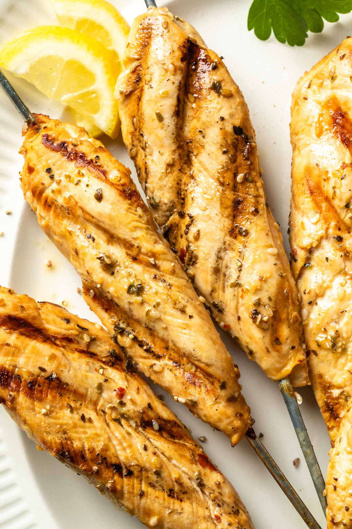 Close up of grilled chicken tenders on metal skewers on a platter with lemon slices and fresh parsley. 