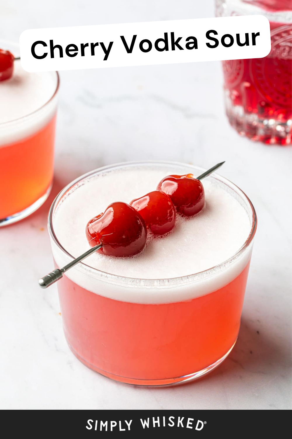 A glass of cherry vodka sour topped with three cherries on a pick. Another similar cherry vodka sour and a red glass are in the background. Text reads “Cherry Vodka Sour” at the top and “Simply Whisked” at the bottom.
