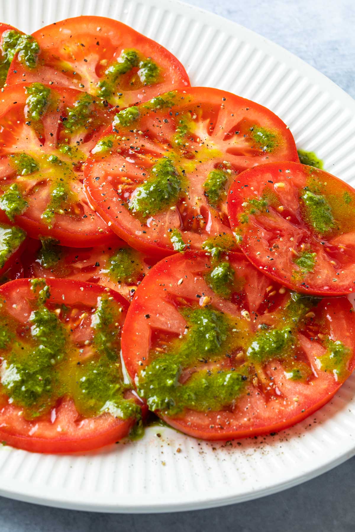Close up of tomatoes sliced on a platter and dressed with basil vinaigrette and salt and pepper. 