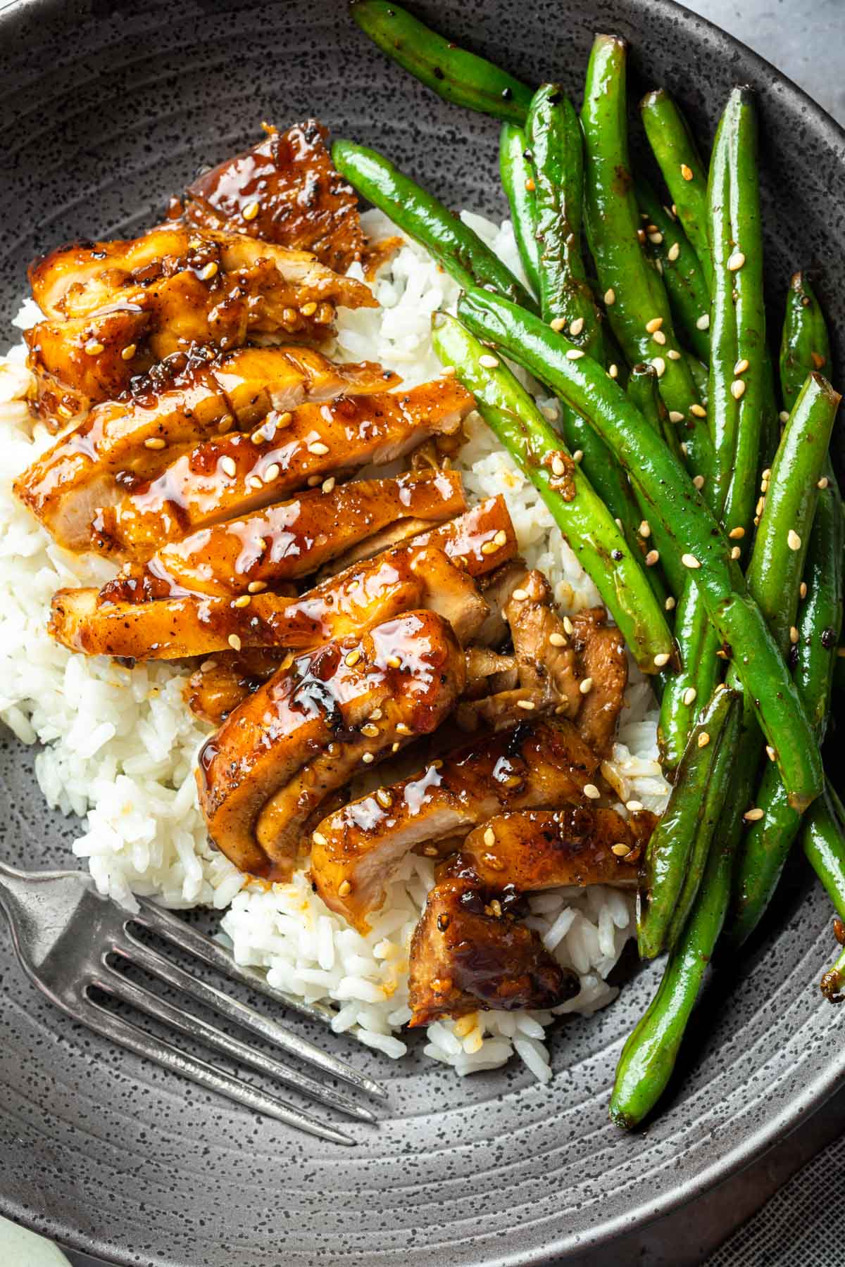 A close up photo of a bowl of white rice topped with teriyaki chicken and green beans, garnished with sesame seeds. 