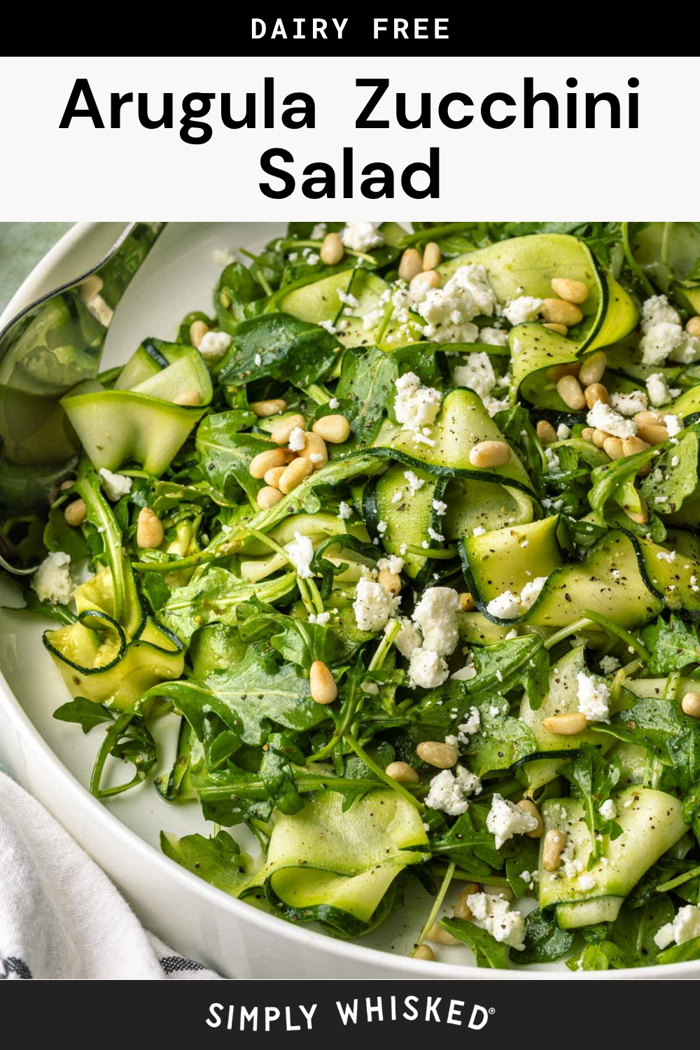 A bowl of fresh zucchini salad featuring arugula, pine nuts, and crumbled cheese, elegantly garnished with cracked pepper. A serving spoon rests in the bowl. Text above reads “Arugula Zucchini Salad” and below reads “Simply Whisked.”.