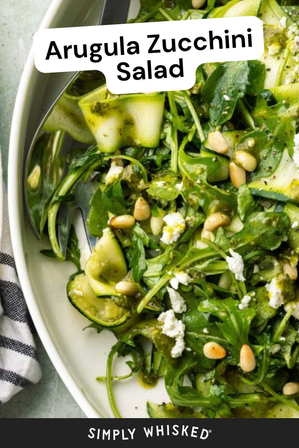 A close-up of zucchini salad featuring arugula, pine nuts, and crumbled cheese on a white plate, served with a fork and spoon. Text reads Arugula Zucchini Salad and Simply Whisked.