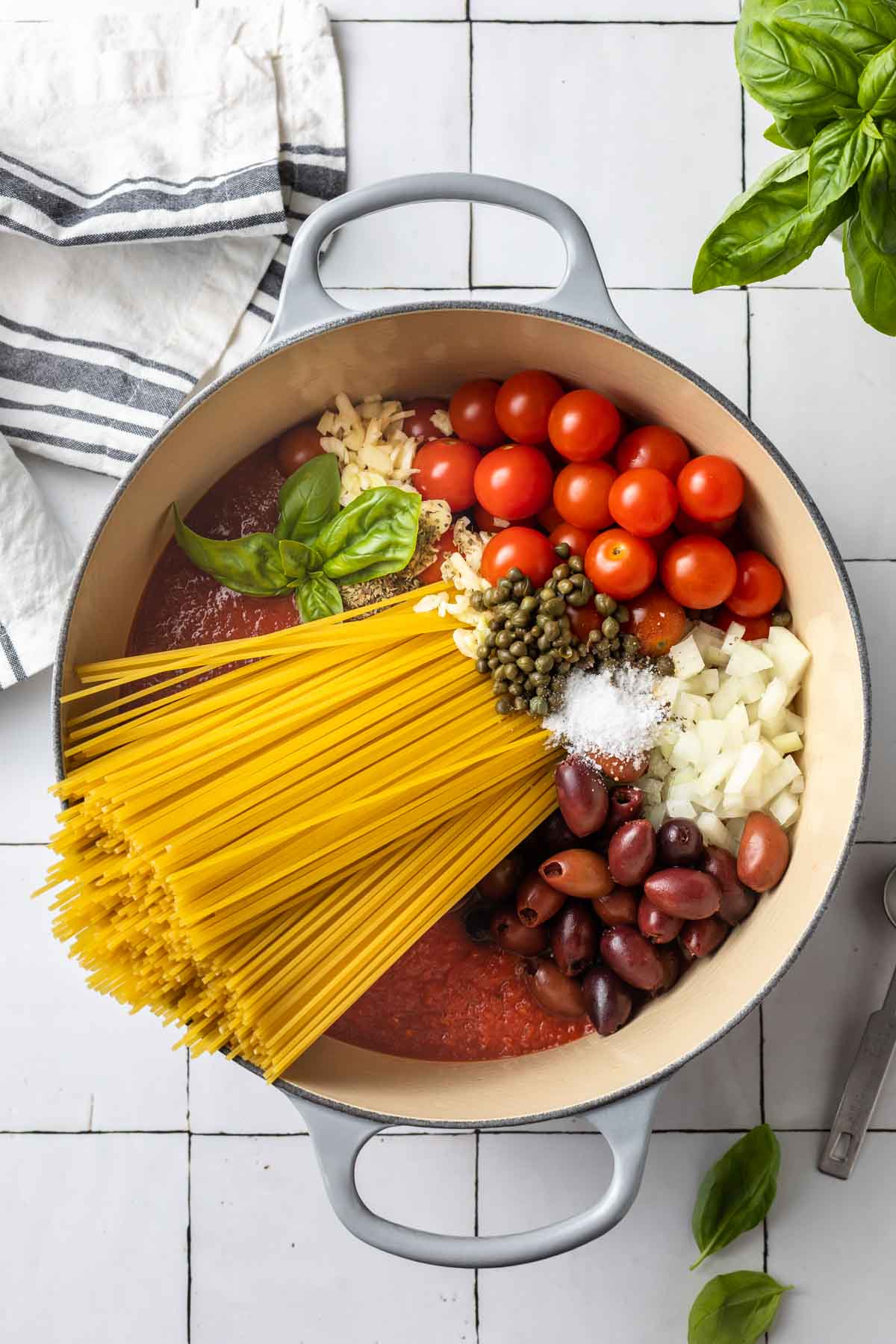 Large stockpot with spaghetti, tomato sauce, capers, onions, cherry tomatoes, kalamata olives, basil leaves, garlic, and salt