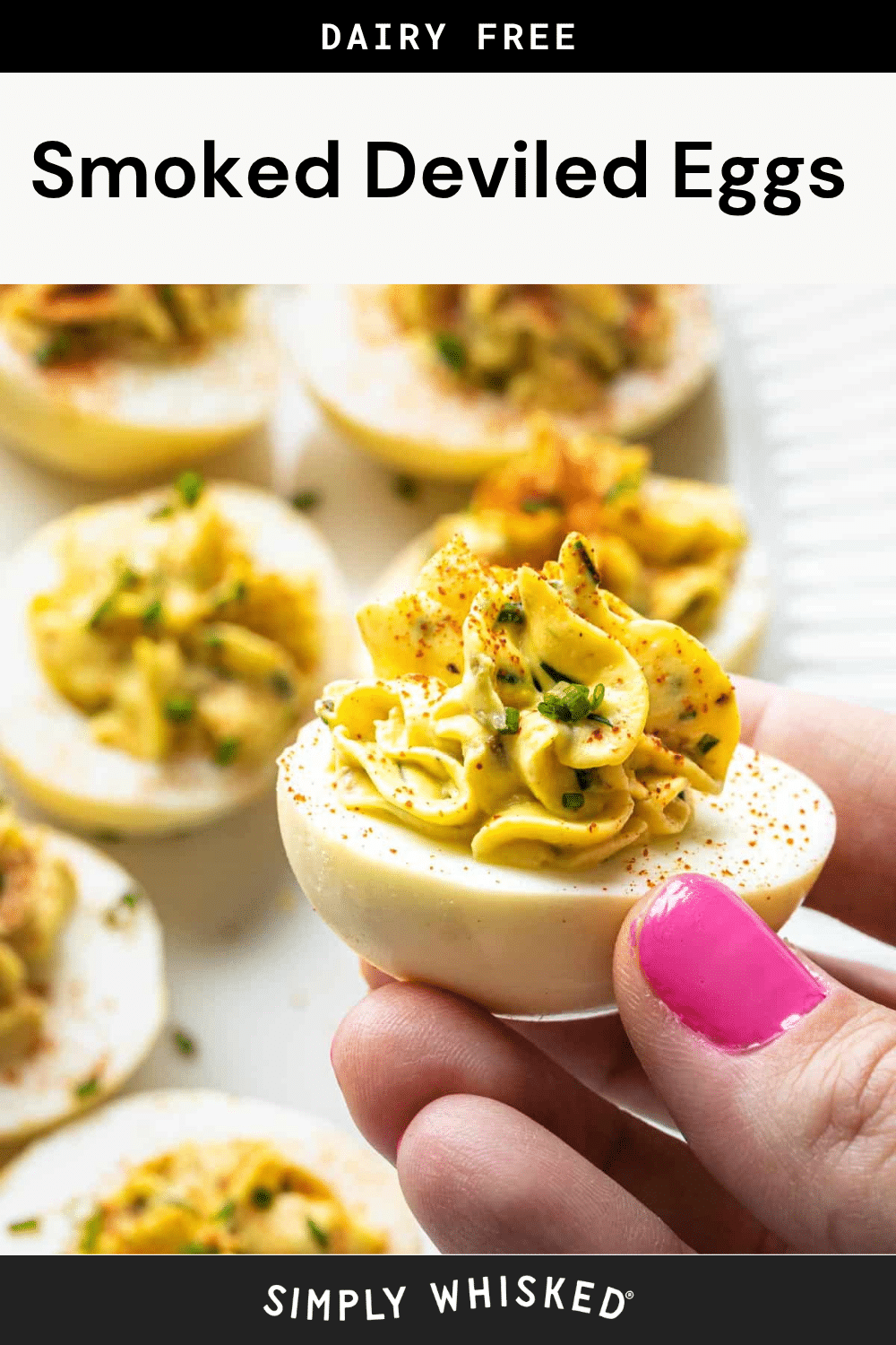 A hand with pink nail polish holds a perfectly seasoned smoked deviled egg topped with chives, with more smoked deviled eggs blurred in the background. Text above reads Dairy Free Smoked Deviled Eggs.