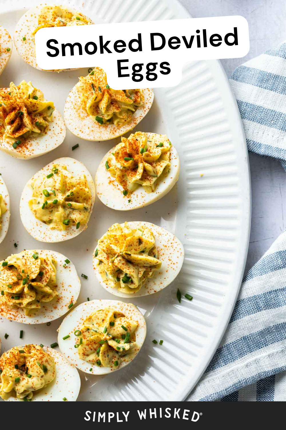 Smoked Deviled Eggs topped with paprika and herbs are arranged on a white platter next to a blue and white striped cloth. The text Smoked Deviled Eggs is at the top, and Simply Whisked is at the bottom.