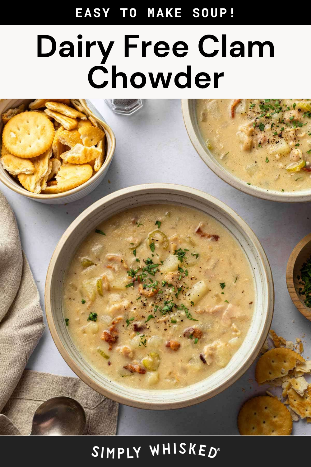Top-down view of Dairy Free Clam Chowder garnished with chopped herbs, next to a bowl of round crackers and a spoon on a napkin. Another bowl of delicious, creamy clam chowder is partially visible in the background.