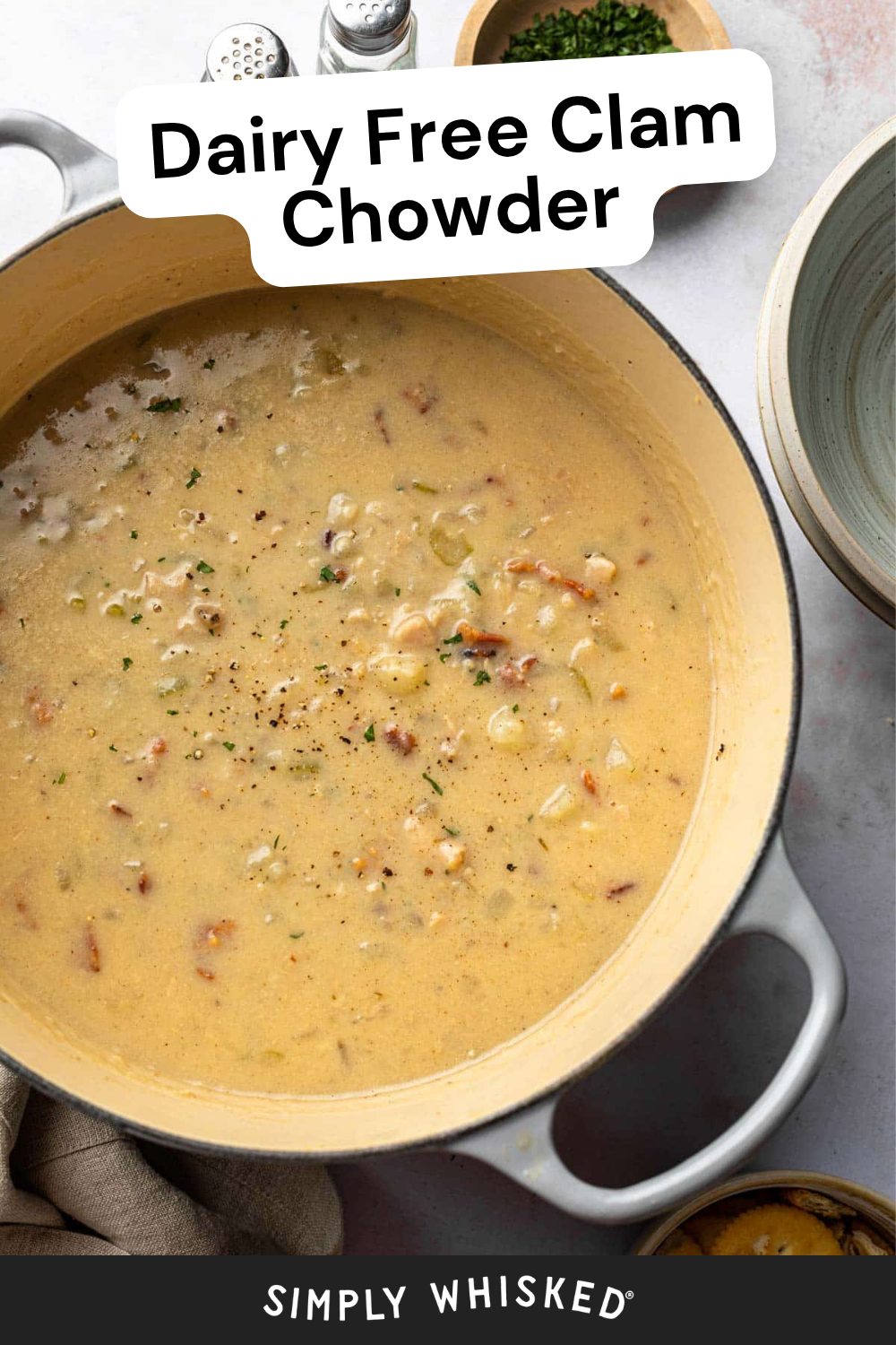 A pot of creamy Dairy Free Clam Chowder garnished with fresh herbs sits on a table beside bowls, salt and pepper shakers, and a dish of chopped chives. Text reads “Dairy Free Clam Chowder.”.