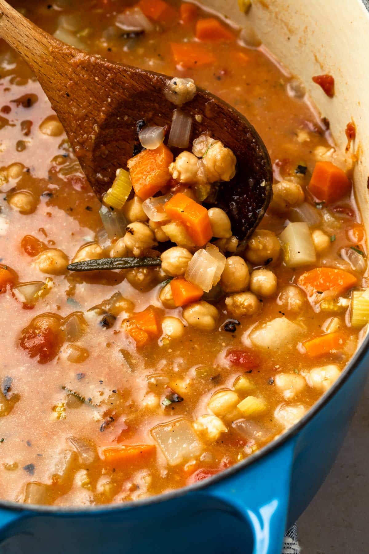 Close up of garbanzo bean soup being lifted by a wooden spoon out of the dutch oven to reveal the ingredients in the soup. 