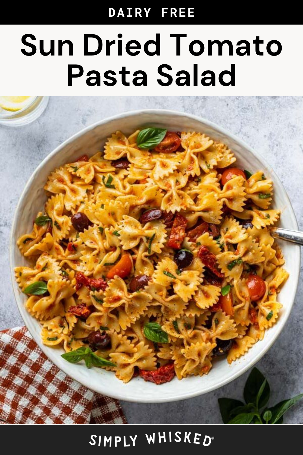 A bowl of Sun Dried Tomato Pasta Salad with farfalle, cherry tomatoes, olives, and fresh basil sits on a light surface, accompanied by a checkered napkin and a small dish of lemon slices.