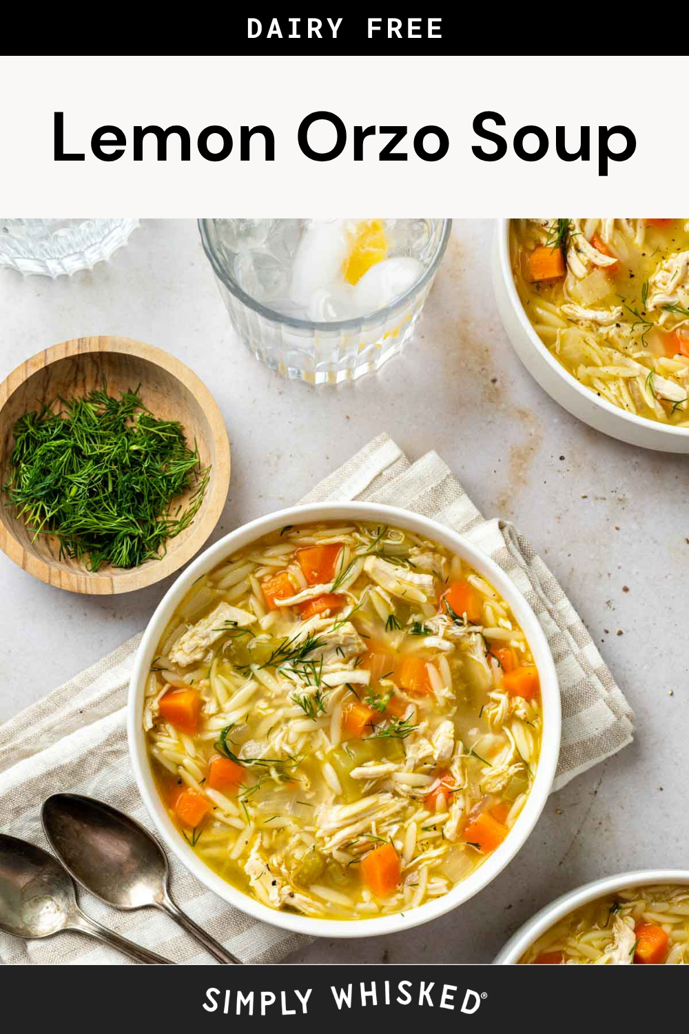 Two bowls of Lemon Orzo Soup with carrots, shredded chicken, and fresh dill sit invitingly on a light table. Spoons, a glass of water with lemon, and a bowl of chopped dill are on the side, all arranged atop a striped napkin for a bright and comforting presentation.