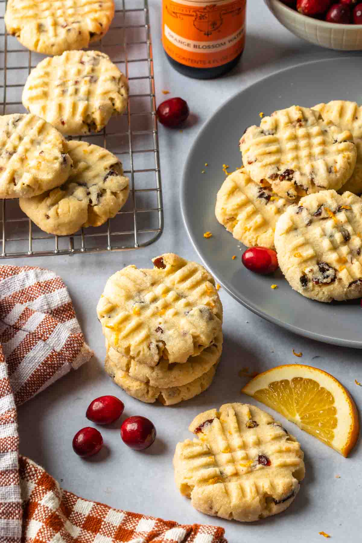 A plate of cookies, next to a cooling rack full of cookies, surrounded by fresh cranberries and orange slices. 
