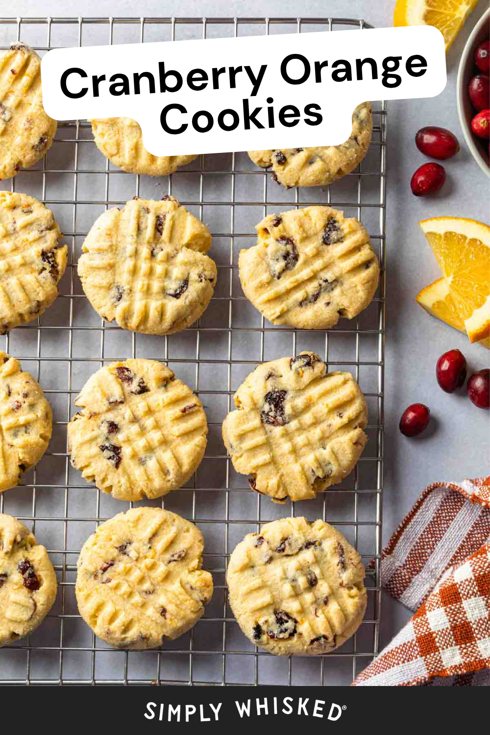 Round Cranberry Orange Cookies cooling on a wire rack, with fresh cranberries and orange slices on the side, and a brown checked napkin partially visible. Text reads Cranberry Orange Cookies, highlighting this delightful blend of citrus and berry flavors.
