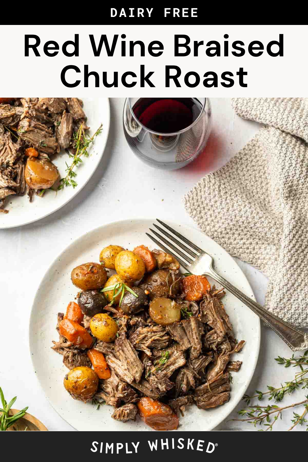 A plate of braised chuck roast with potatoes and carrots, simmered in red wine and garnished with fresh herbs, sits on a white table next to a fork, a beige napkin, and a glass of red wine.