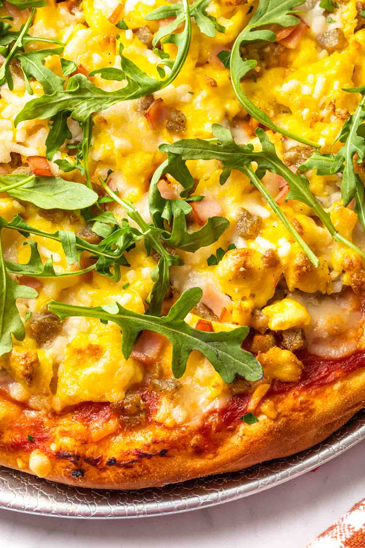 A breakfast pizza topped with scrambled eggs, arugula, canadian bacon and breakfast sausage. 