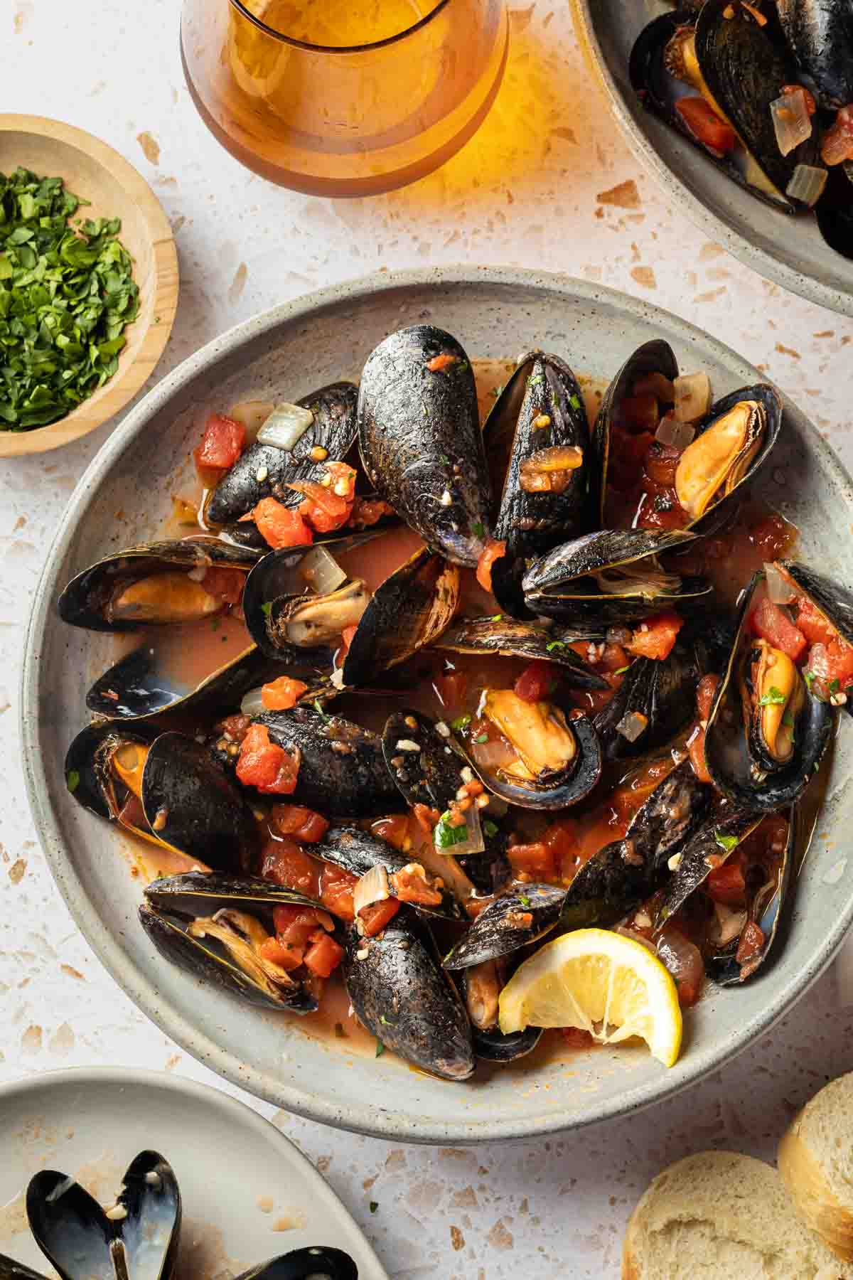 Overhead shot of a bowl of mussels marinara with a lemon wedge, next to some bread slices, and bowl of empty shells and a glass of wine. 
