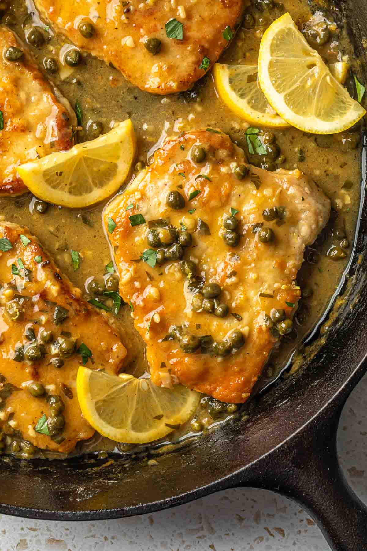 A cast iron skillet full of breaded pork chops with capers and lemon slices. 