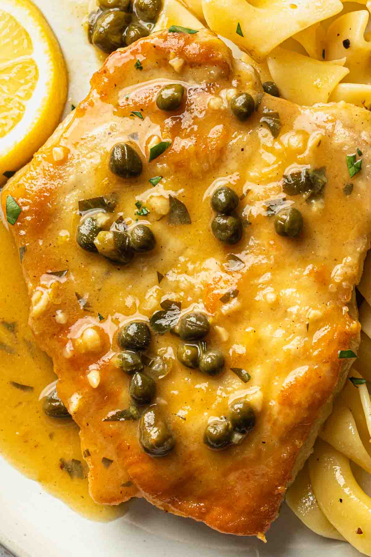 Close up of a breaded pork chop covered in a piccata sauce, showing the capers and fresh herbs in the sauce. 