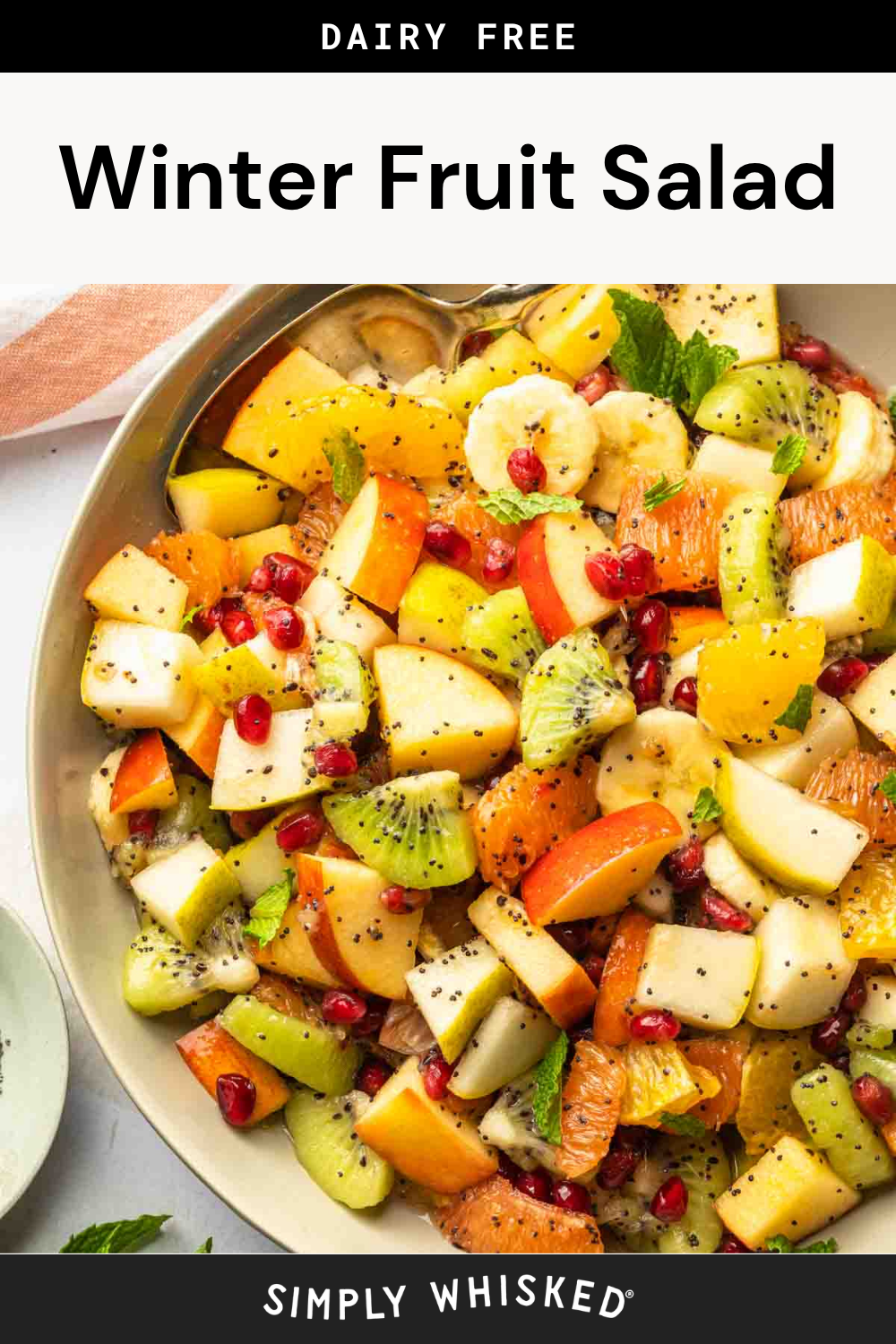 A vibrant bowl of winter fruit salad featuring apples, oranges, bananas, kiwi, pomegranate seeds, and fresh mint leaves. A serving spoon rests in the bowl. The text above reads Dairy Free Winter Fruit Salad.