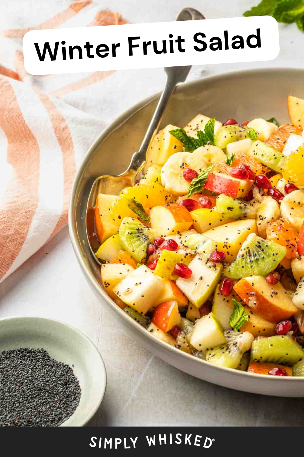 A vibrant bowl of winter fruit salad brims with kiwi, apple, banana, pomegranate seeds, and fresh mint, all sprinkled with poppy seeds. A spoon rests in the bowl, ready to enjoy this colorful winter fruit salad, while a small dish of extra poppy seeds sits alongside.