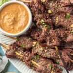 A plate of grilled beef satay skewers garnished with chopped herbs and peanuts, served with a bowl of creamy peanut dipping sauce. A small dish of fresh herbs sits nearby on the table.