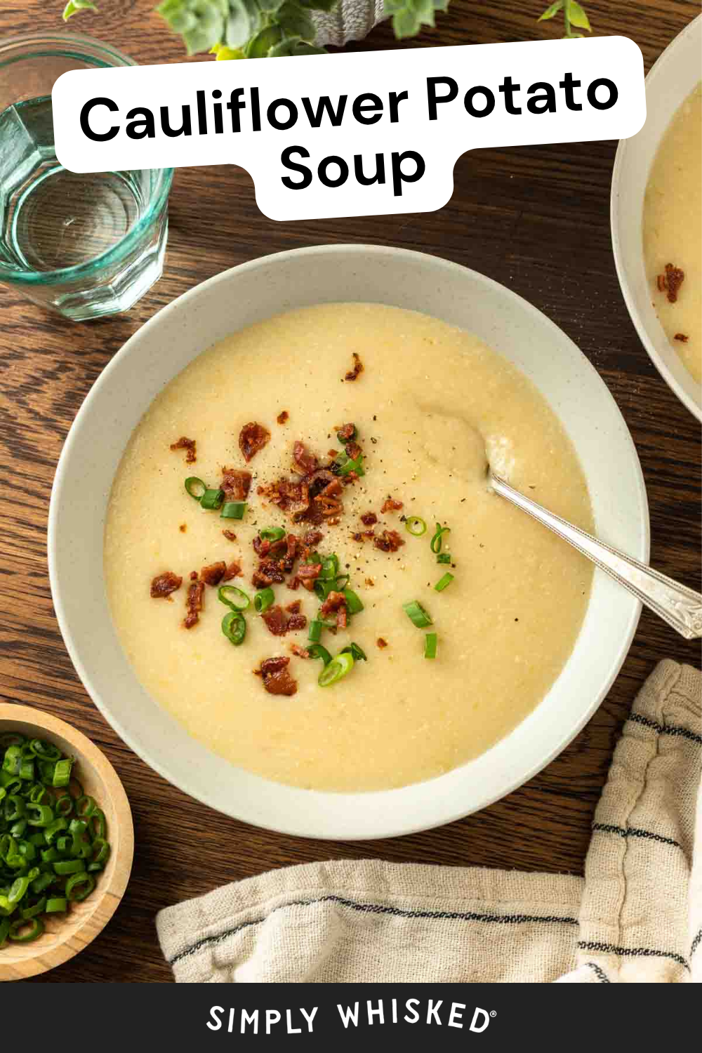 A bowl of creamy Cauliflower Potato Soup topped with chopped green onions and bacon bits, with a spoon, a bowl of green onions, water glass, and striped napkin nearby.