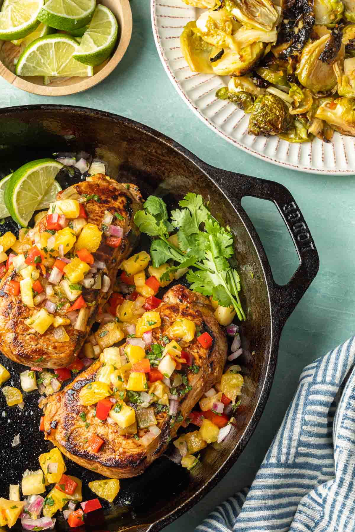 Two pineapple pork chops covered with pineapple salsa in a cast iron skillet next to a platter of brussels sprouts. 