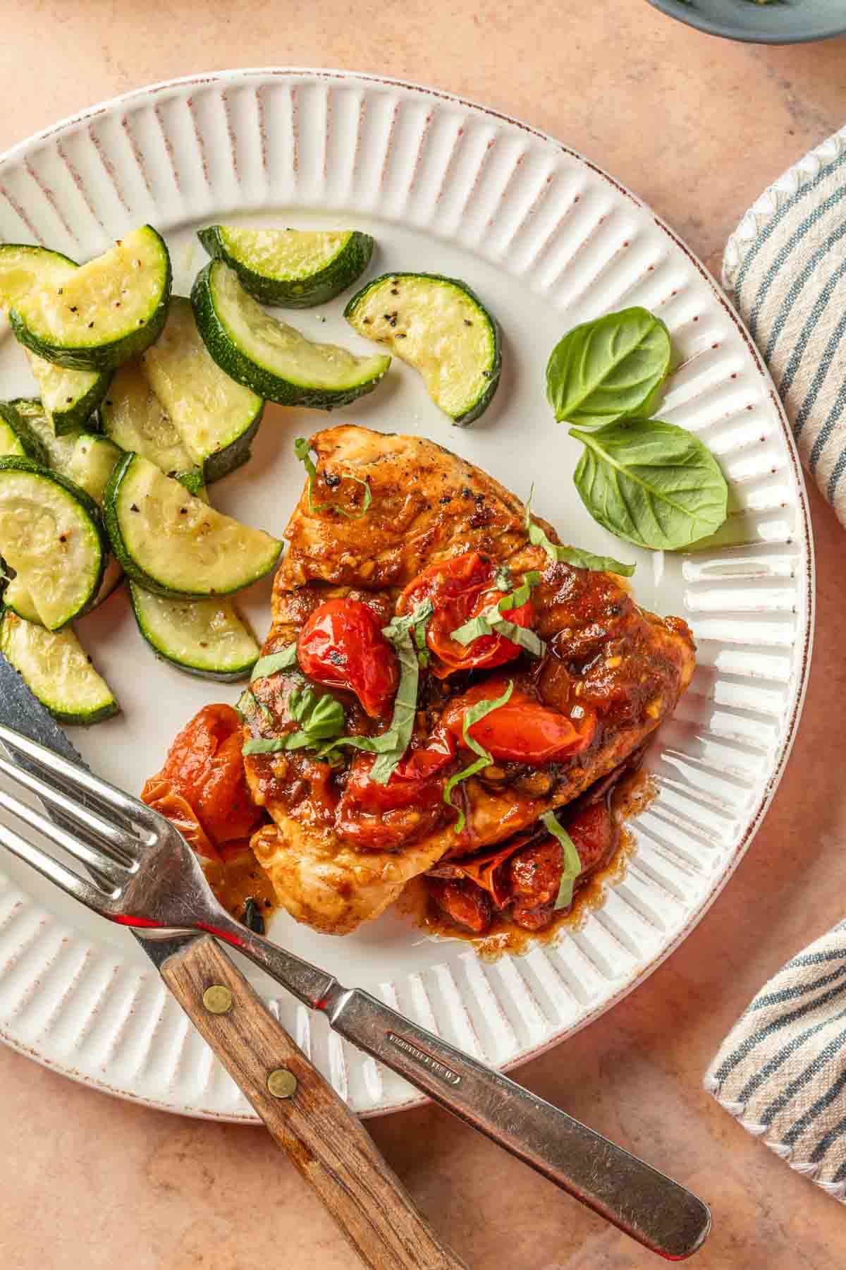 Close up of chicken covered in a roasted tomato sauce, on a plate with roasted zucchini slices. 