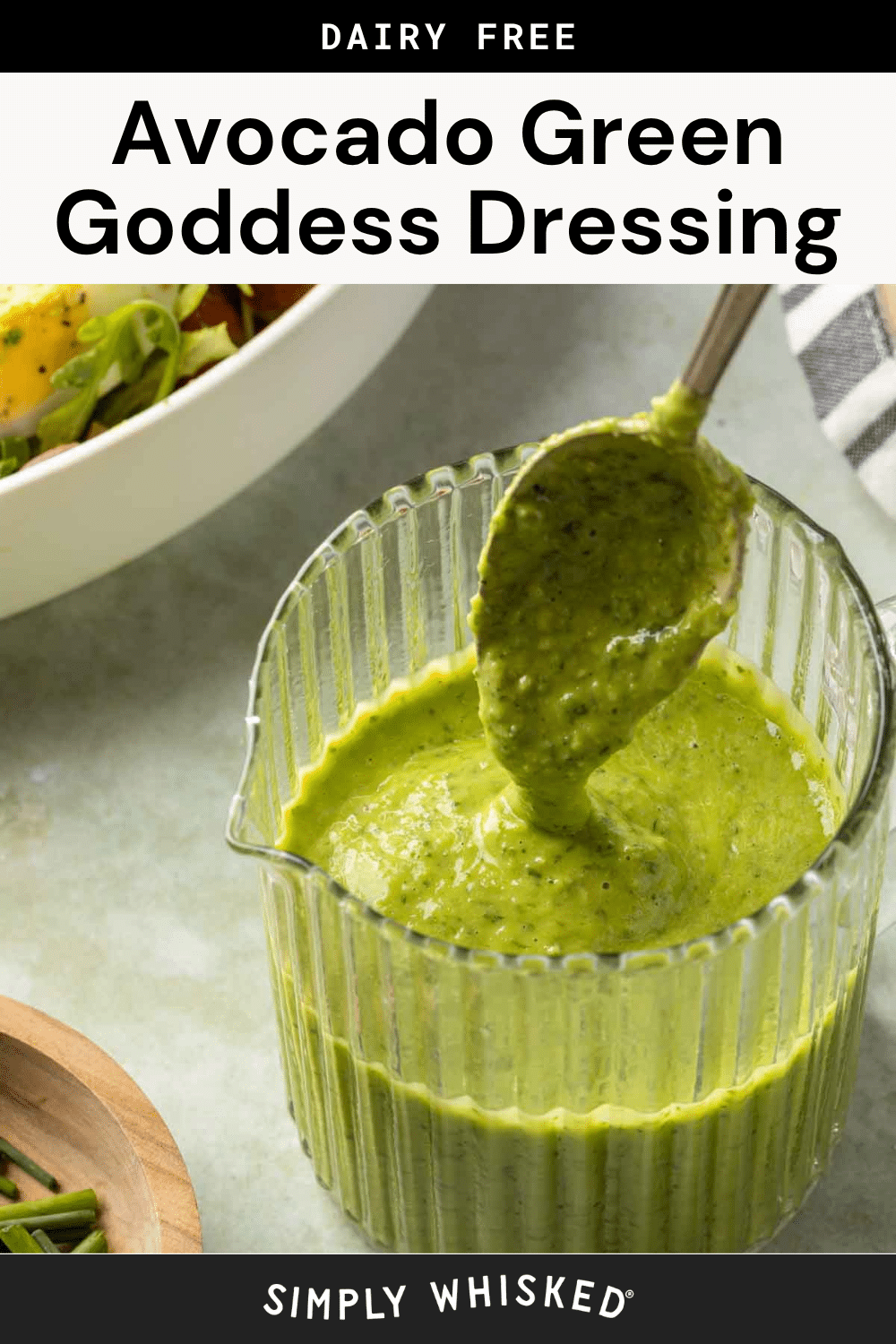 A glass measuring cup filled with creamy Avocado Green Goddess Dressing, with a spoon lifting some out. In the background, there’s a salad bowl and a striped napkin. Text reads Dairy Free Avocado Green Goddess Dressing.