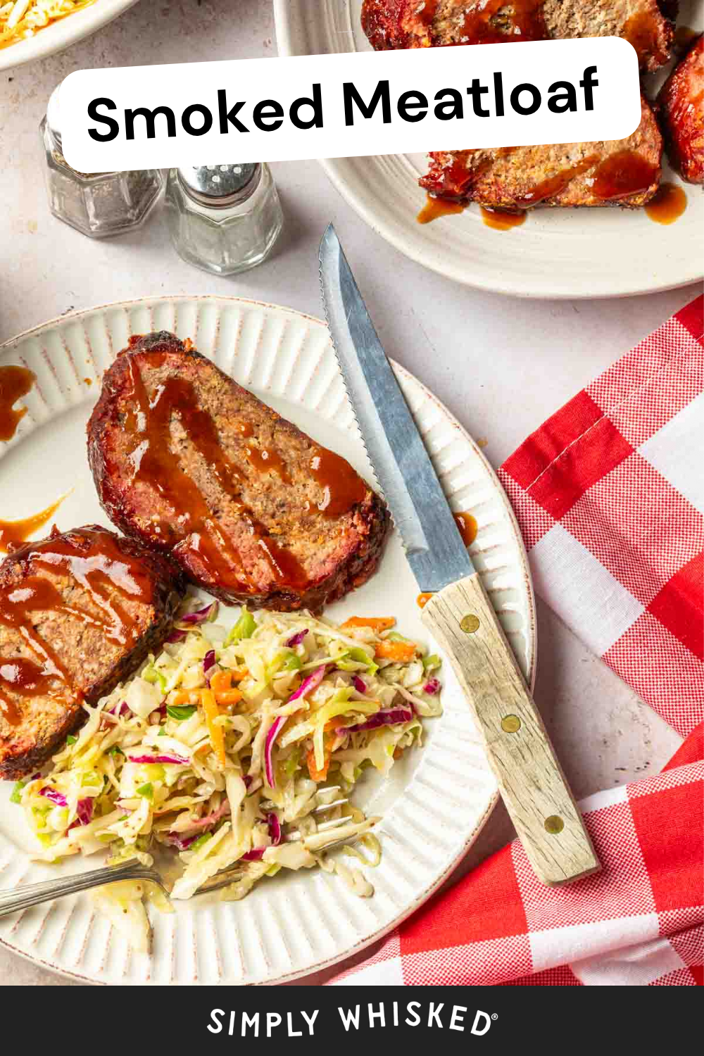 Two slices of smoked meatloaf topped with sauce are served on a white plate with creamy coleslaw, complete with a fork and knife, red checkered napkin, and salt and pepper shakers nearby.