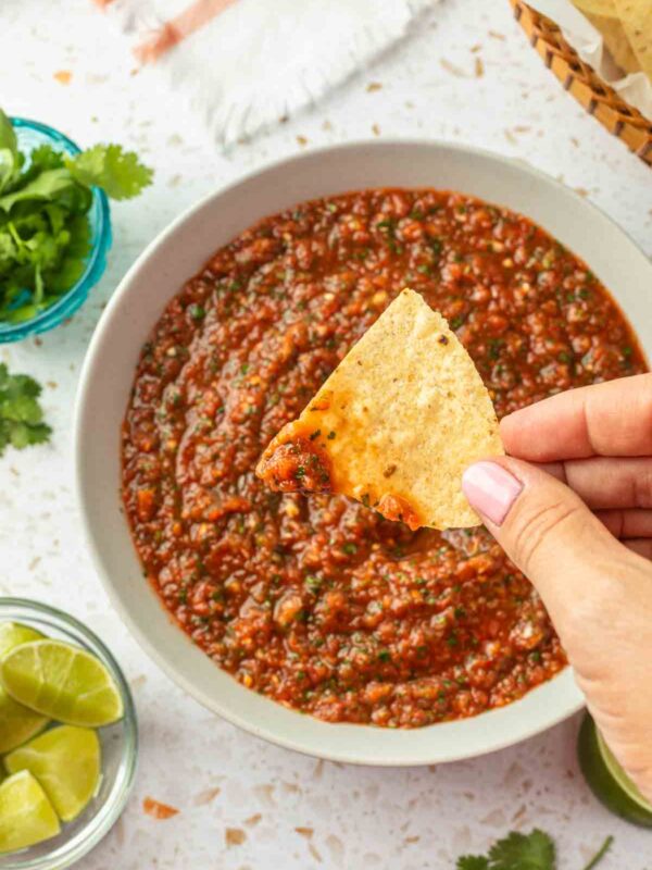 A hand holding a tortilla chip dipped in salsa, over the bowl of salsa.