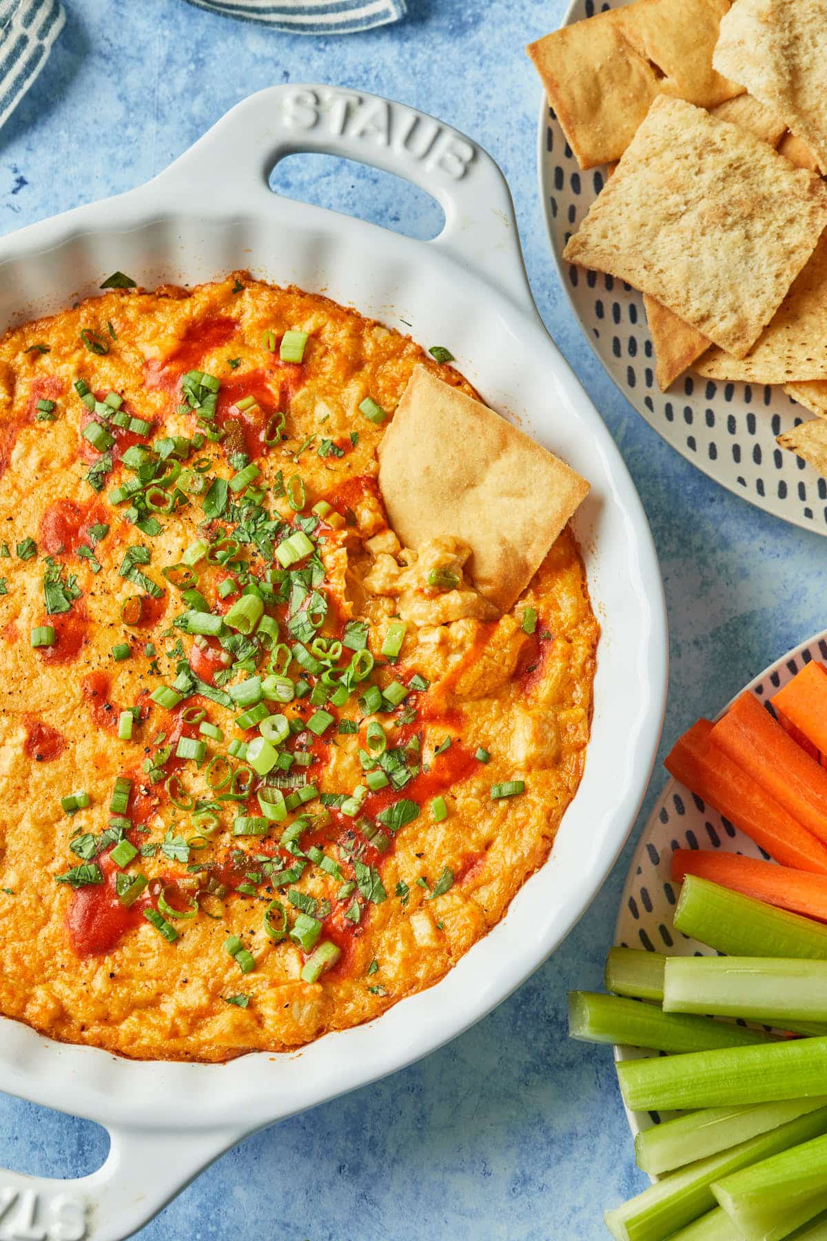A pie plate of healthy buffalo chicken dip, garnished with green onions and a drizzle of buffalo sauce, with a pita chip dipped into the side. 