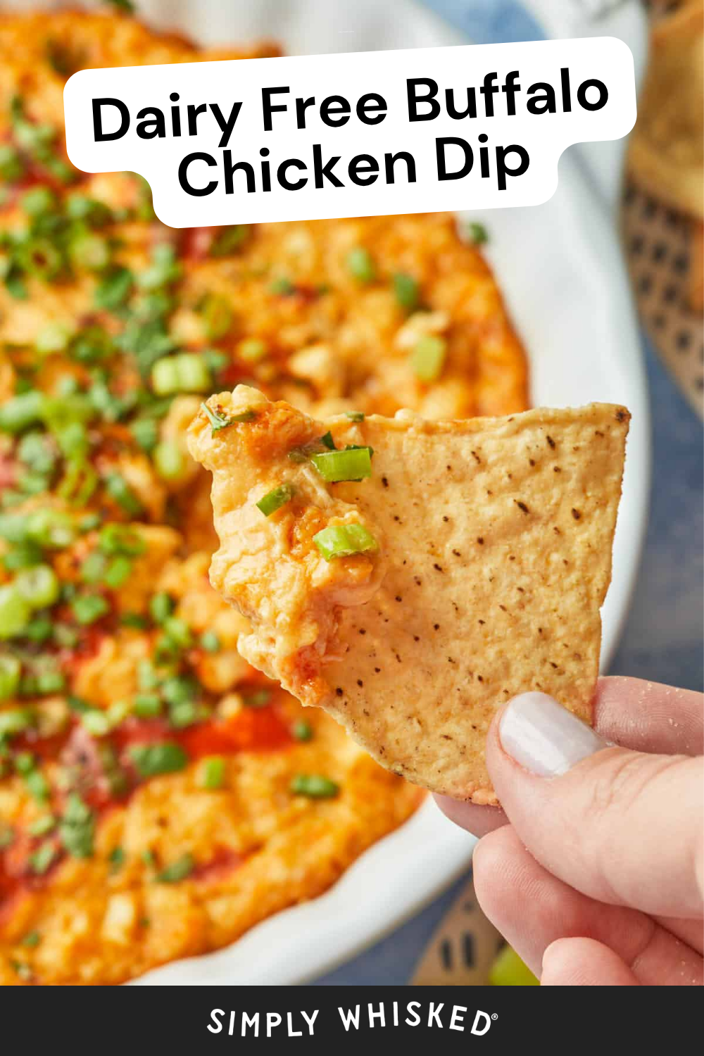 A hand holds a tortilla chip with healthy buffalo chicken dip, topped with chopped green onions, above a dish filled with the dip. Text reads Dairy Free Buffalo Chicken Dip.