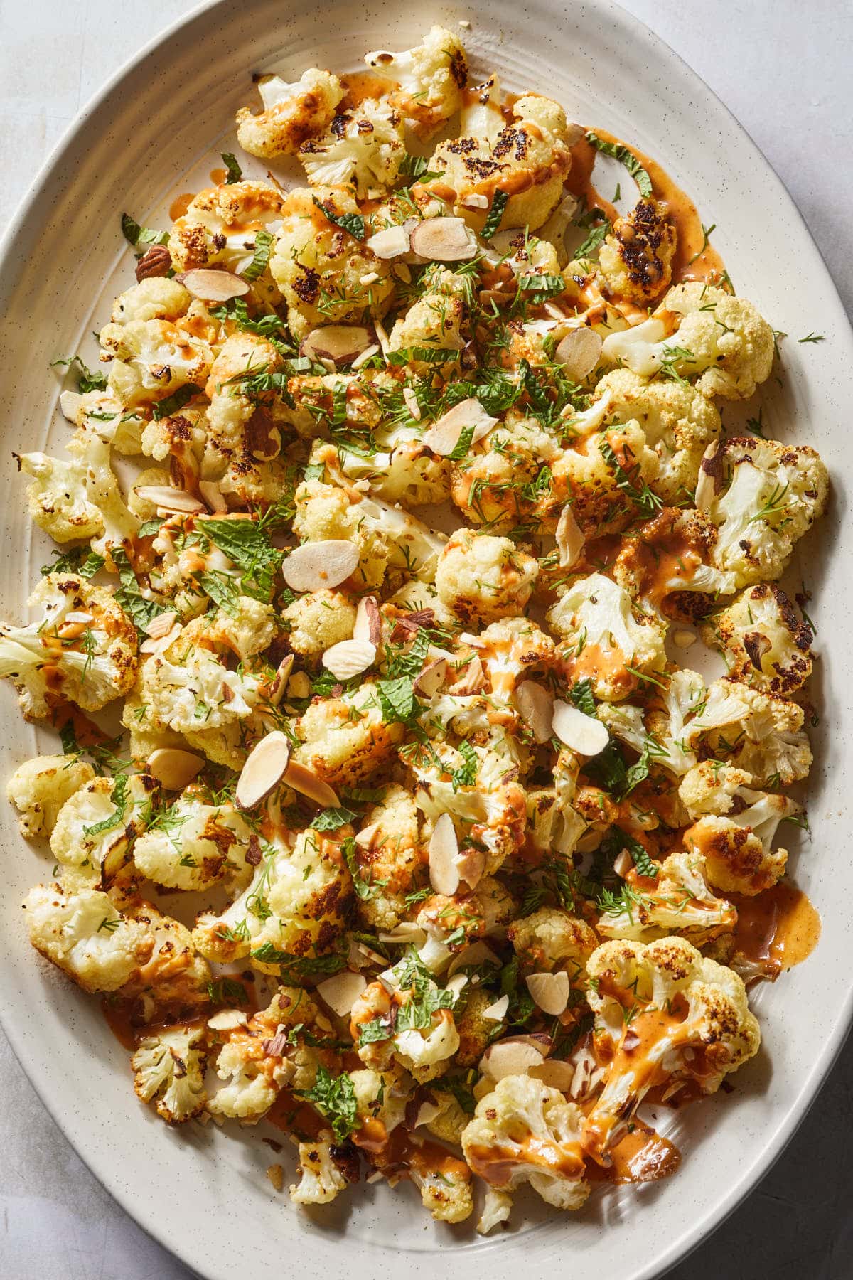 A serving platter of roasted veggies topped with tahini, harissa, fresh herbs and slivered almonds. 