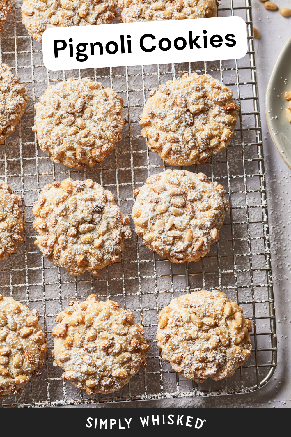 Pignoli Cookies dusted with powdered sugar rest on a wire cooling rack, with a plate and a few pine nuts visible in the corner, highlighting the classic elegance of these Italian treats.
