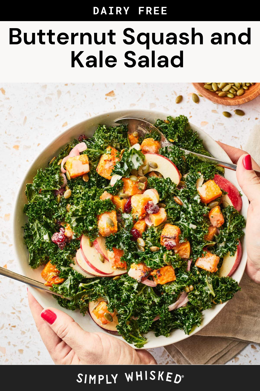 A large bowl of Butternut Squash and Kale Salad brimming with apple slices, cranberries, and seeds sits ready to enjoy. Two hands with red nails cradle the bowl, while a fork and scattered seeds rest nearby on a speckled surface.