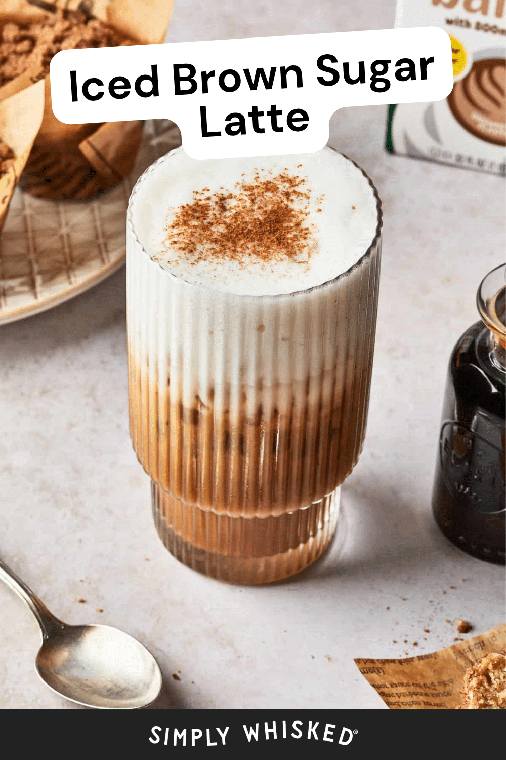 A glass of iced brown sugar latte topped with creamy foam and sprinkled cinnamon sits on a marble surface, surrounded by a spoon, a small bottle of syrup, and brown sugar. Text reads refreshing brown sugar latte.