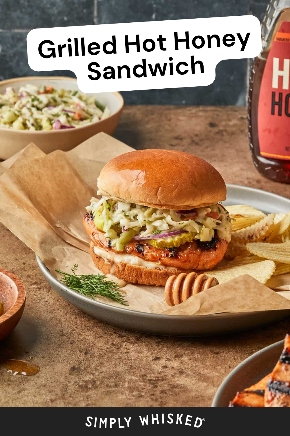 A Hot Honey Chicken Sandwich topped with coleslaw sits on parchment paper with potato chips, a honey dipper, and fresh dill. A bottle of hot honey and bowls of slaw are in the background. Text reads Grilled Hot Honey Sandwich.