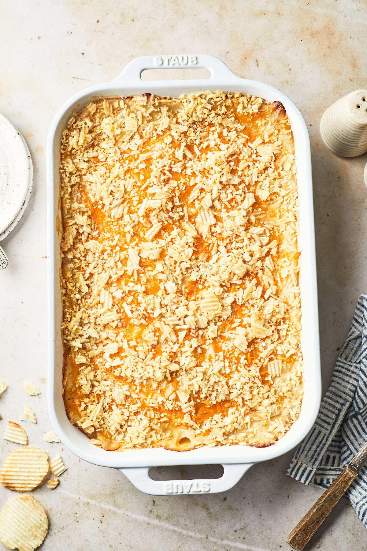 A white Staub baking dish filled with dairy-free cheesy potato casserole, topped with a layer of crushed potato chips for a crispy texture. The dish sits on a light-colored countertop with some potato chip crumbs scattered around.