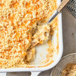 A close-up of a partially scooped dairy-free cheesy potato casserole in a white baking dish, showing a creamy and golden interior. A serving spoon rests in the dish, highlighting the creamy and cheesy texture under the crunchy chip topping.