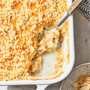 A close-up of a partially scooped dairy-free cheesy potato casserole in a white baking dish, showing a creamy and golden interior. A serving spoon rests in the dish, highlighting the creamy and cheesy texture under the crunchy chip topping.