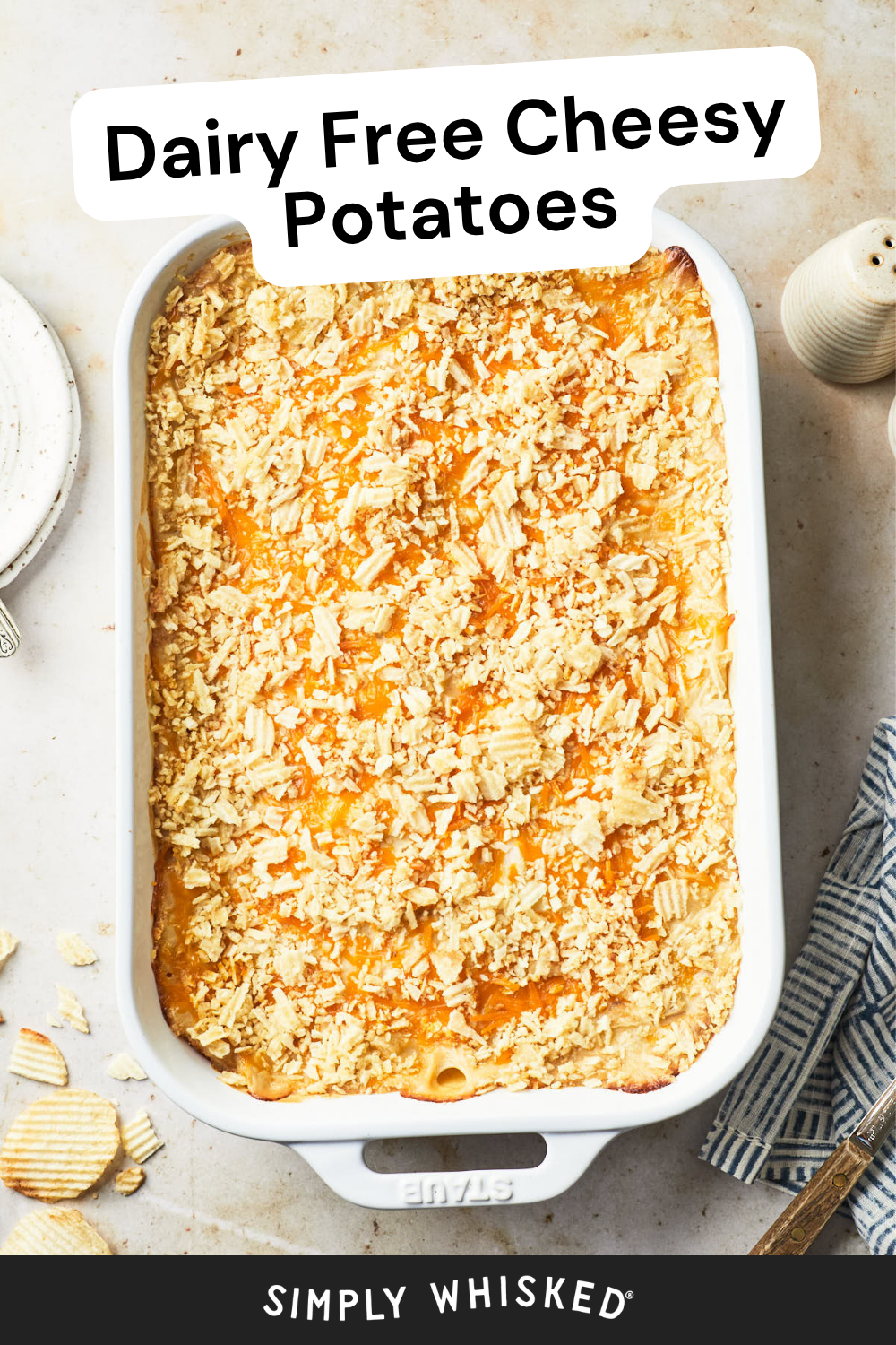 A white baking dish filled with Cheesy Potatoes, made dairy free and topped with crushed chips. Text reads Dairy Free Cheesy Potatoes. Some plates, a fork, and potato chips are nearby.