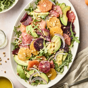A large oval plate features an avocado citrus salad of vibrant citrus slices, creamy avocado, arugula, red onion, and pine nuts. Nearby are a bowl of arugula, fresh oranges, dressing, and salt and pepper shakers on a light table.