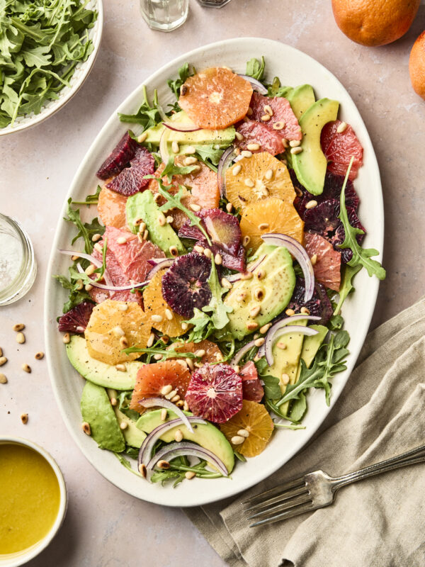A large oval plate features an avocado citrus salad of vibrant citrus slices, creamy avocado, arugula, red onion, and pine nuts. Nearby are a bowl of arugula, fresh oranges, dressing, and salt and pepper shakers on a light table.