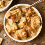 A bowl of creamy French onion soup topped with toasted bread cubes and melted cheese, garnished with fresh thyme, with a spoon in the bowl and another dish partially visible in the background.