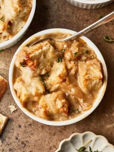 A bowl of creamy French onion soup topped with toasted bread cubes and melted cheese, garnished with fresh thyme, with a spoon in the bowl and another dish partially visible in the background.